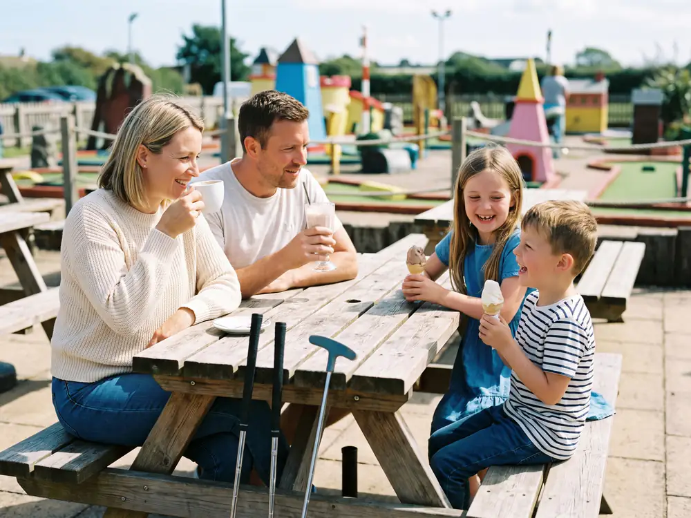 A family enjoys ice creams, a milkshake and a coffee at a picnic table beside the course, their putters leaning on the table