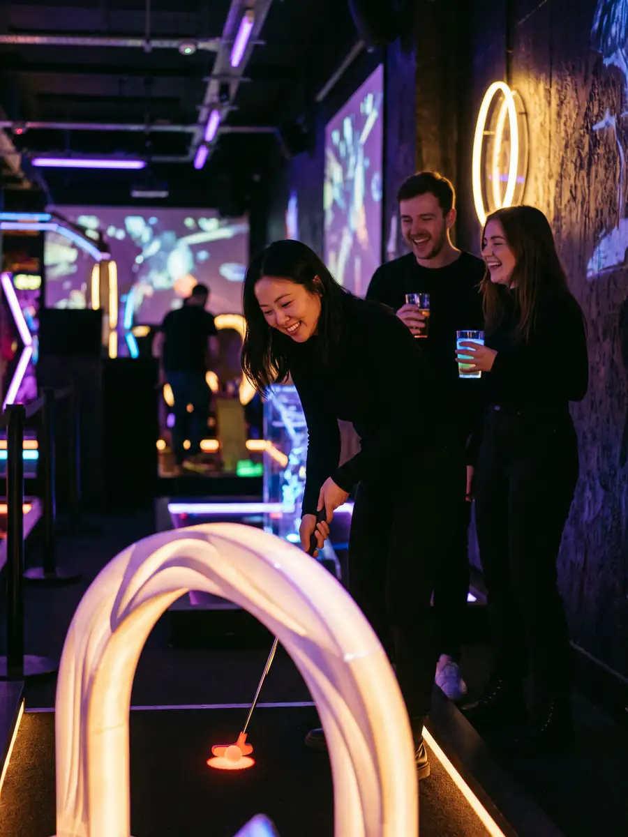 A woman putts through a glowing arch on an indoor neon course while two friends with drinks laugh behind her