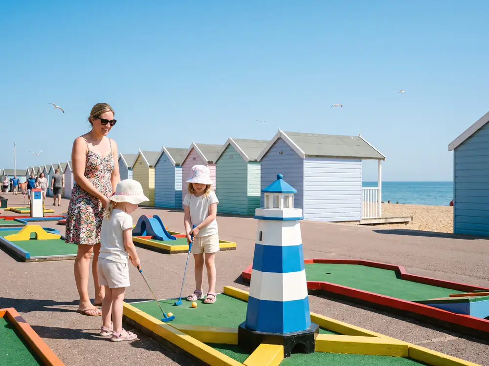 Two little girls in sun hats putt beside a blue and white lighthouse on a seafront course, with pastel beach huts and the sea behind