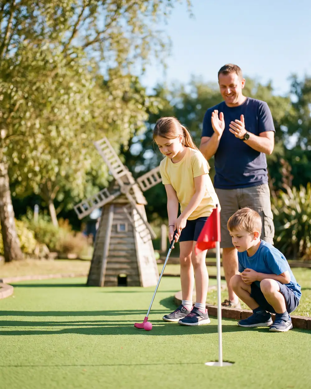 A girl in a yellow T-shirt putts a pink ball towards a red flag while her little brother crouches to watch and her dad claps, with a wooden windmill behind them