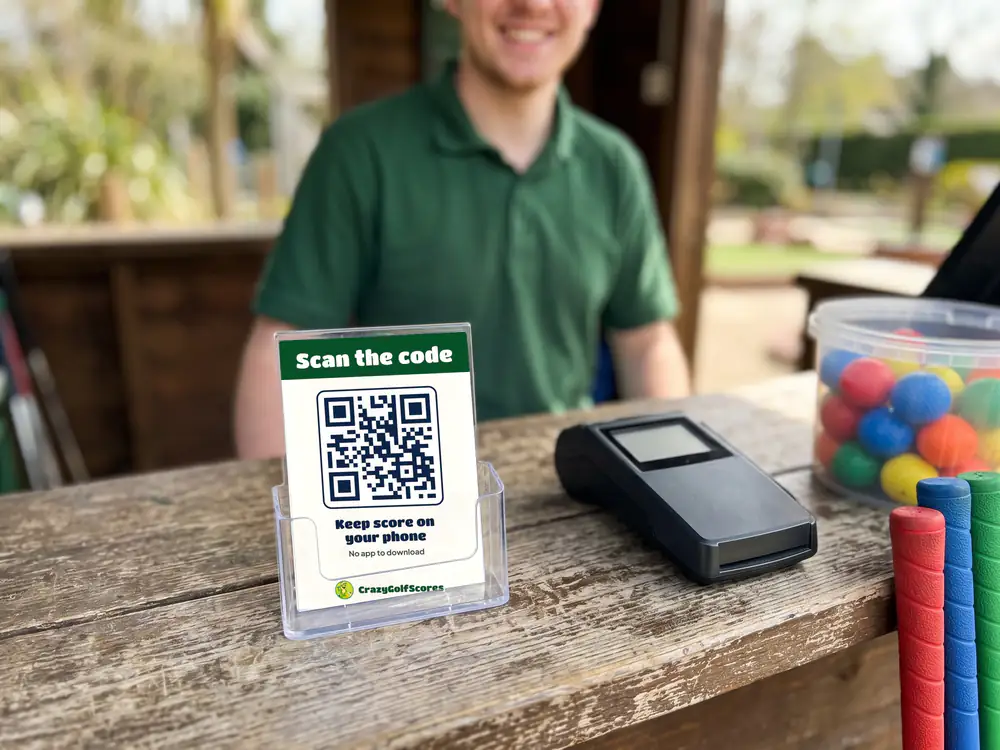 A Scan the code card with a QR code in a clear stand on the kiosk counter, beside the card reader and a tub of golf balls