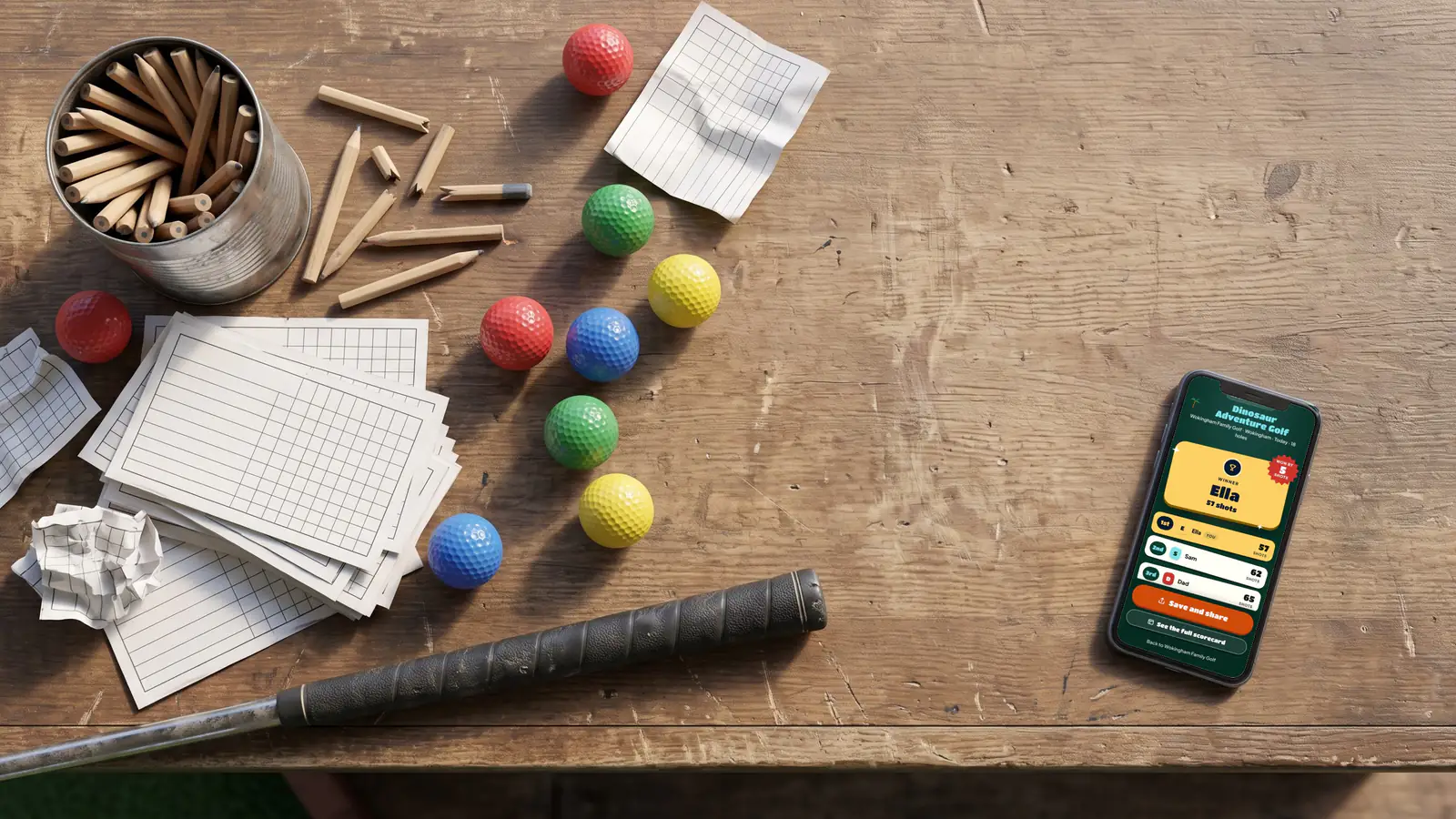 A kiosk counter with a tin of stubby pencils, a stack of blank paper scorecards and coloured balls, and a phone beside them showing the winner of a round