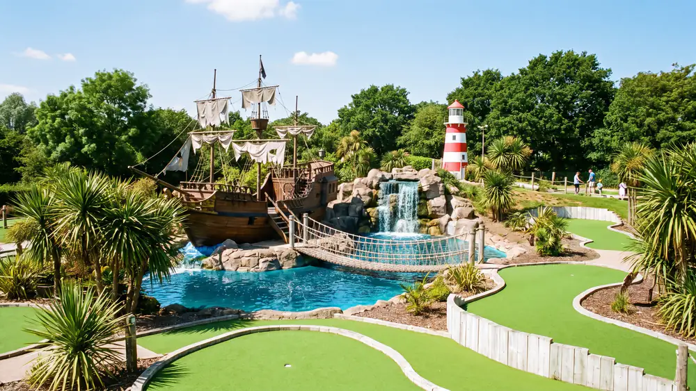 An adventure golf course from above: a wooden pirate ship, a waterfall into a blue pool, a rope bridge and a red and white lighthouse among palms