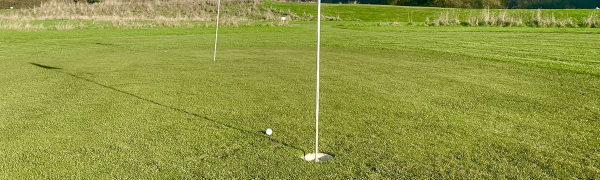 Two flagsticks on a wide green in low sun, with a ball a few feet from the near cup