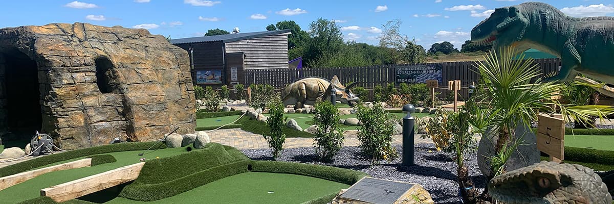Two greens on the dinosaur course with a ball and a putter on one, the rock face and cave on the left and a triceratops and a tyrannosaurus among the planting