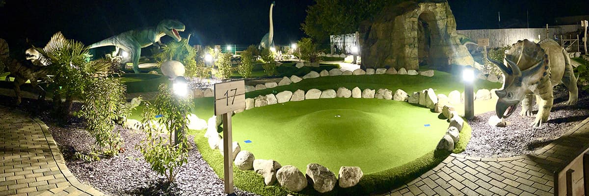 The dinosaur course lit at night: a green under floodlights, the marker for hole 17, the rock arch behind and dinosaurs standing in the dark