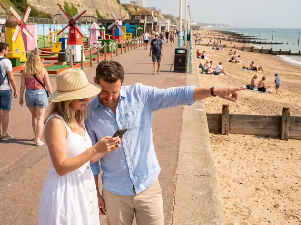 A couple on a seaside promenade look at a phone and point ahead to a colourful crazy golf course with windmills
