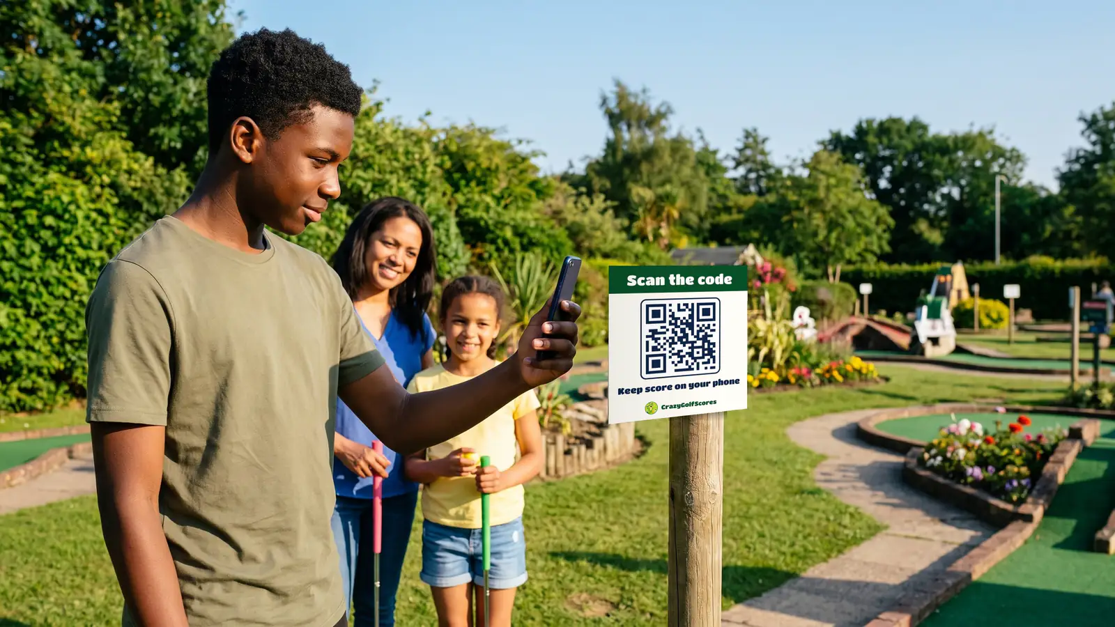 A teenage boy points his phone at a Scan the code sign on a post by the first hole, while his mum and sister wait with their putters